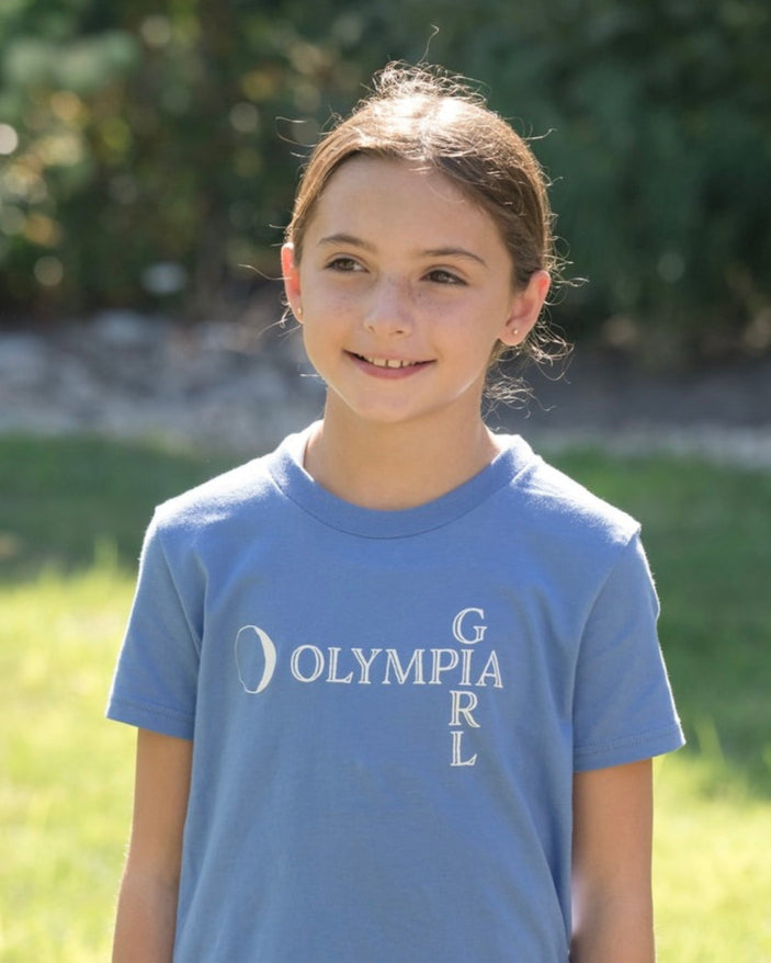Young girl wearing a blue t-shirt with 'Olympia Girl' text outdoors.