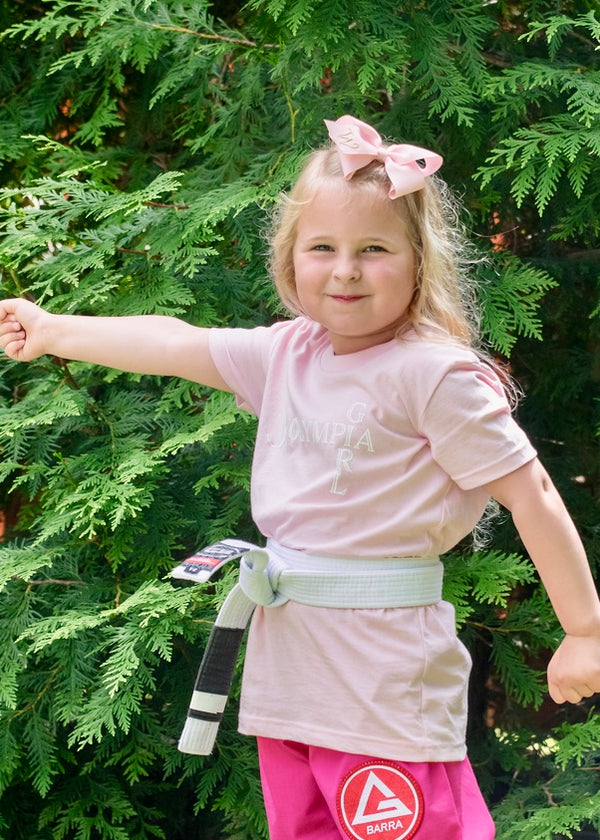 Child in pink shirt and pink shorts with a belt, standing in front of green trees