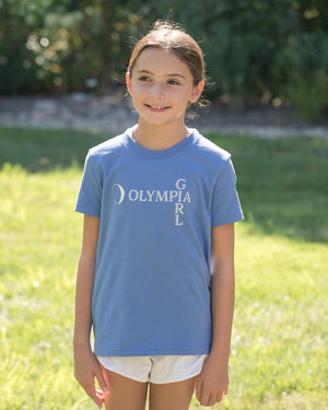 Young girl wearing a blue t-shirt with 'Olympia Girl' text, standing outdoors on grass.