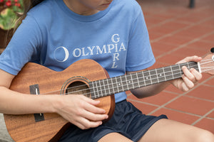 Person playing a ukulele wearing a blue t-shirt with 'Olympia Girl' text.